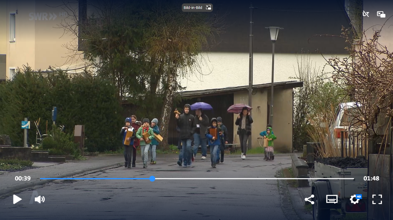Screenshot aus dem Video. Man sieht eine Gruppe Kinder, begleitet von zwei Erwachsenen, mit ihren Klappern bei Regen auf einer Straße laufen.
