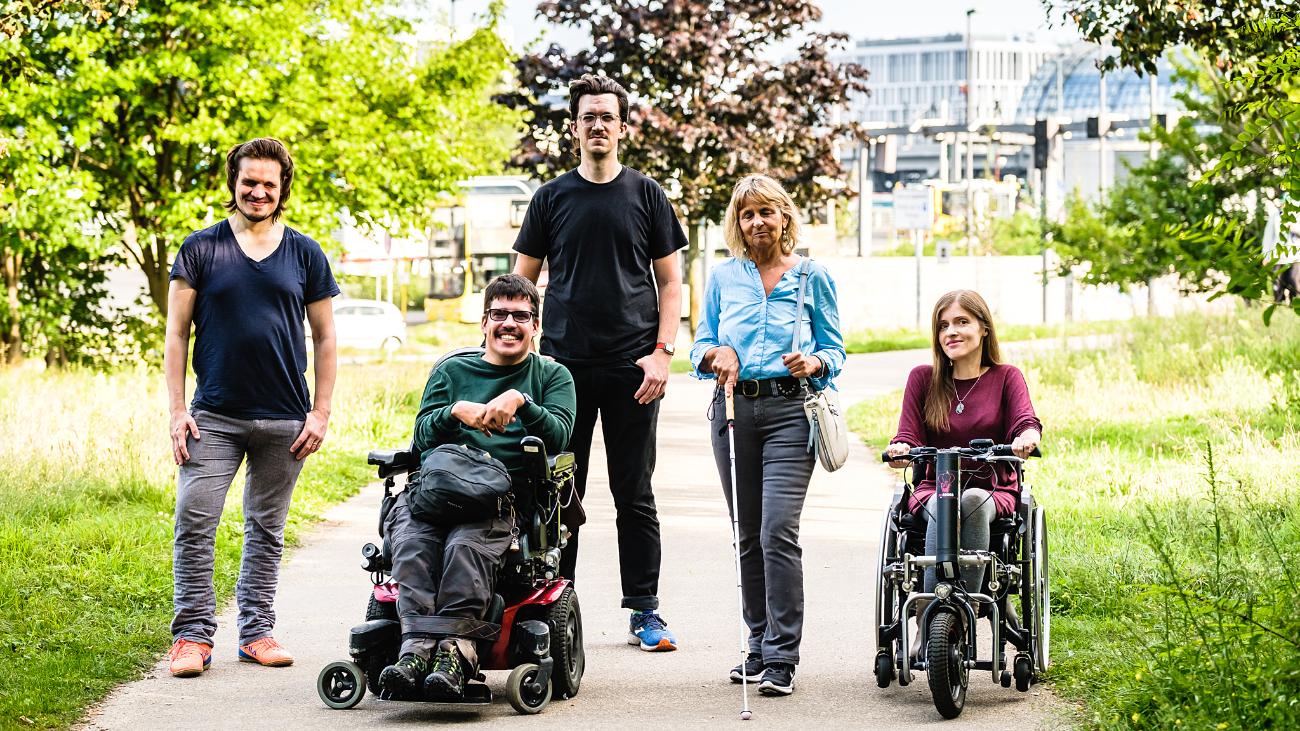 Fünf Personen mittleren Alters, zwei Personen nutzen elektrische Rollstühle, eine Frau hält einen weißen Blindenstock, zwei junge Männer stehen im Hintergrund