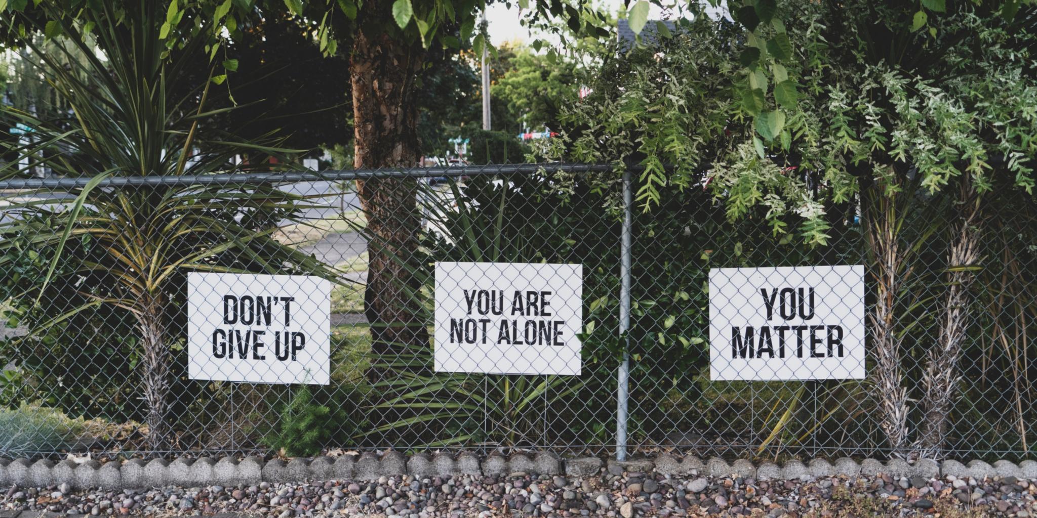 Man sieht einen Maschendrahtzaun, dahinter drei Schilder in Englisch beschriftet. Übersetzt: Geben Sie nicht auf! Sie sind nicht allein! Sie sind wichtig!
