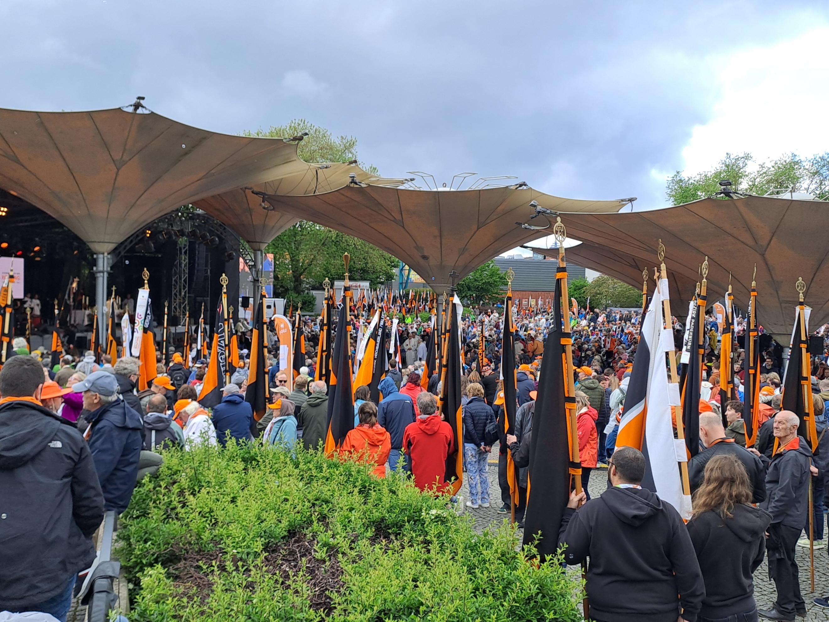 Auf dem Bild ist eine große Menschenmenge bei einer Veranstaltung im Freien zu sehen, viele von ihnen tragen orangefarbene Kleidung oder Accessoires. Zahlreiche orange-schwarze Fahnen mit goldenen Spitzen werden aufrecht gehalten. Im Hintergrund befindet sich eine überdachte Bühne mit Lautsprechern und Bildschirmen unter großen, zeltartigen Dächern. (Alt-Text erstellt mit Unterstützung von ChatGPT)