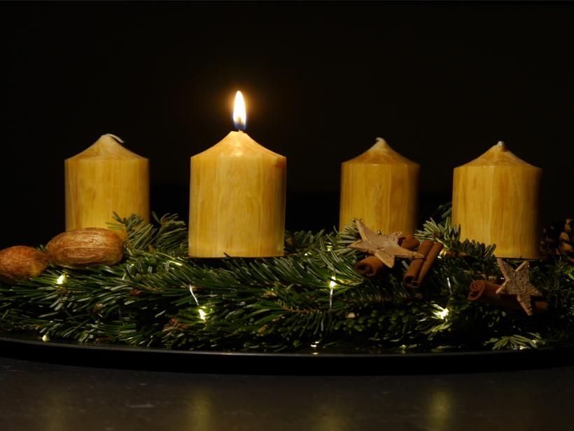 Das Foto zeigt in Großaufnahme einen Adventskranz vor dunklem Hintergrund. Der Adventskranz hat honigfarbene Kerzen, von denen eine brennt.