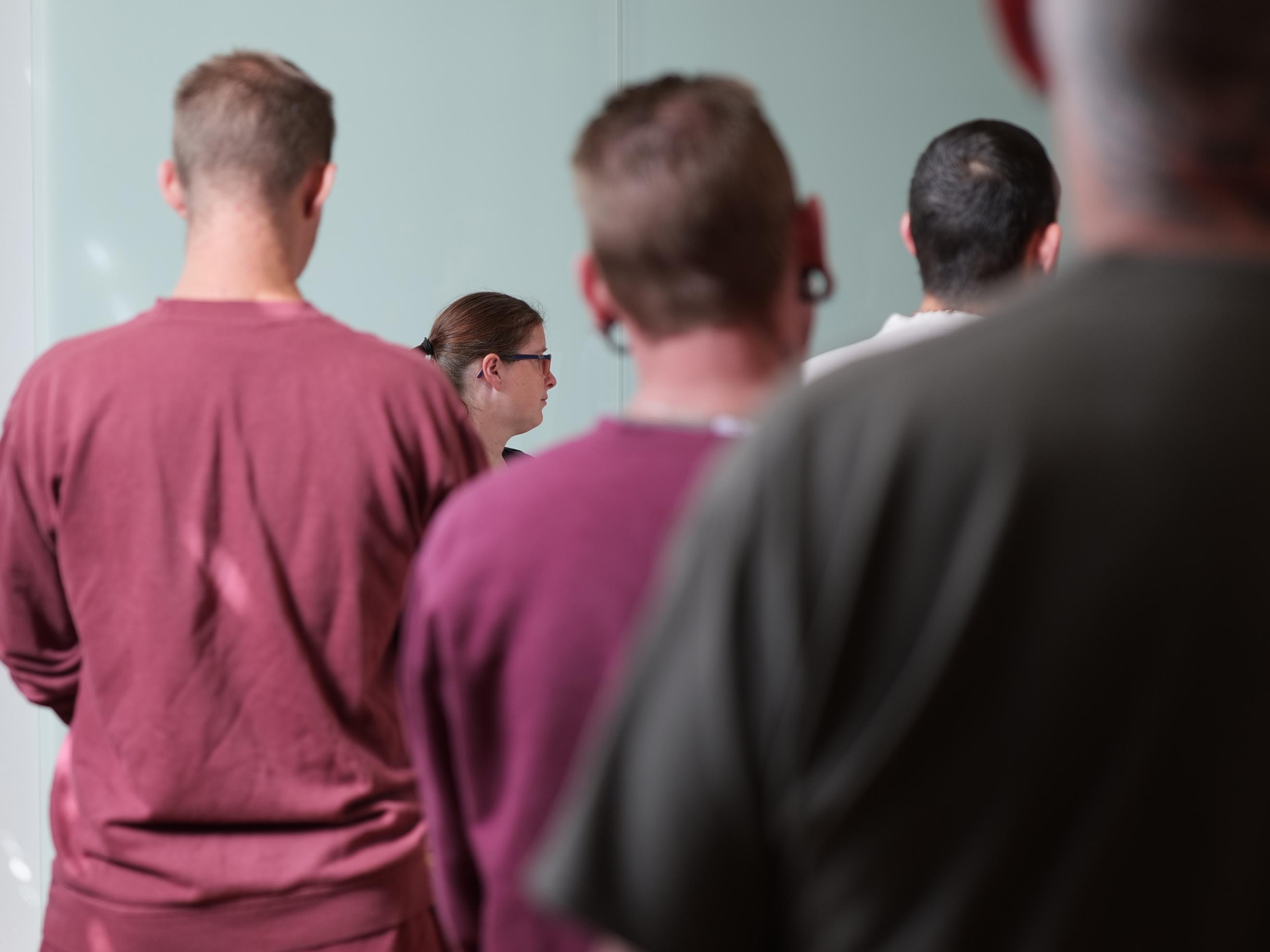 Das Foto zeigt – teilweise unscharf - Oberkörper und Köpfe von vier Männern von hinten, zum Teil verdecken sie sich gegenseitig. Etwas im Hintergrund, zwischen zwei der Männer, der Kopf einer Frau im Profil; es handelt sich dabei um Clara Boor, die Hauptperson der Podcast-Folge. Über das Foto ist in dünnen weißen Großbuchstaben der Satz gelegt: „Clara Boor ist Organistin in der JVA“.