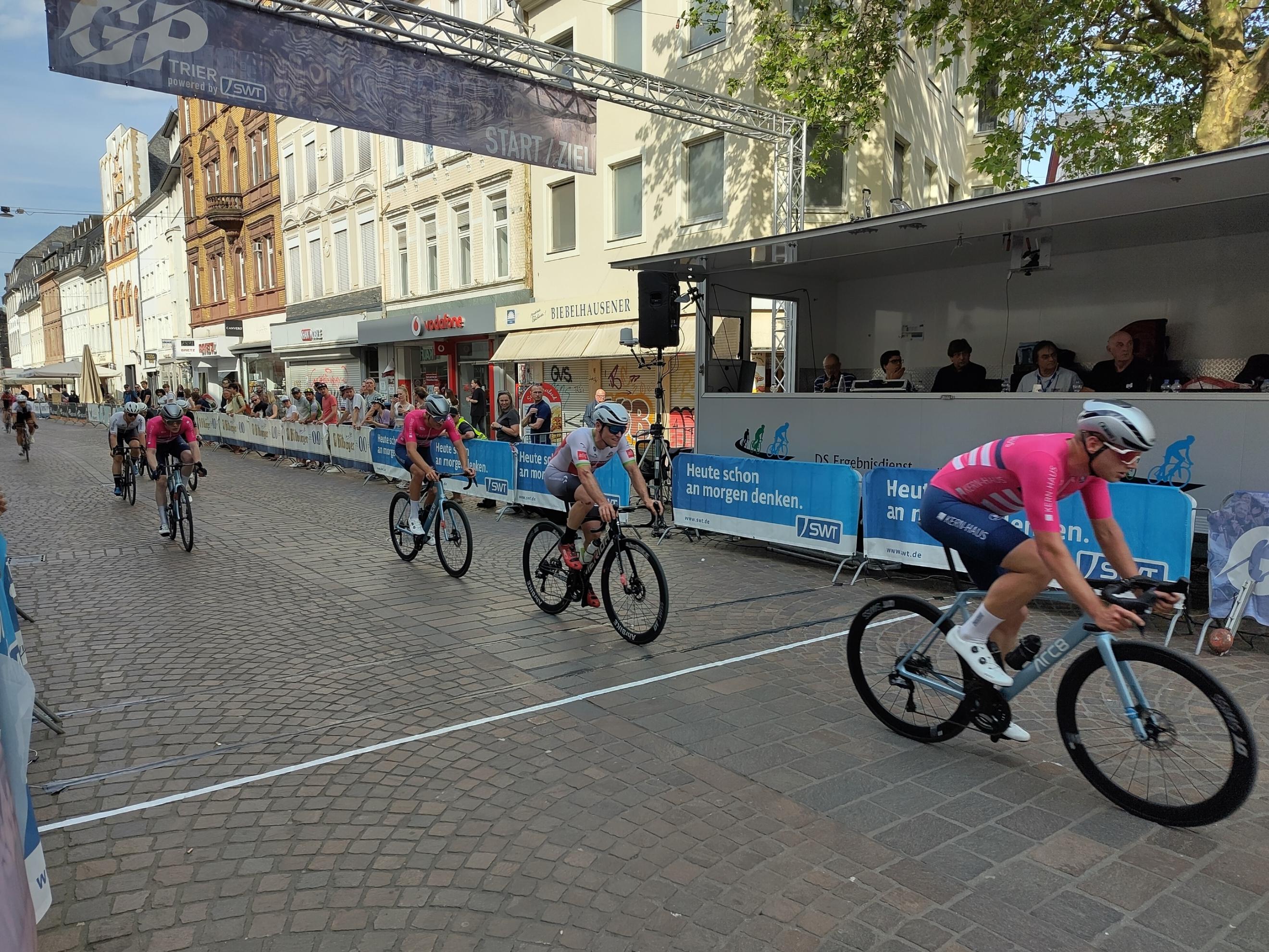 Foto eines Radrennens. Auf einer abgesperrten Innenstadtstraße (es handelt sich um einen Abschnitt der Trierer Fußgängerzone) fährt ein halbes Dutzend Radsportler von links nach rechts durch Bild, weiter hinten folgen weitere Fahrer. Hinter den Absperrungen am Rand stehen Zuschauer*innen.