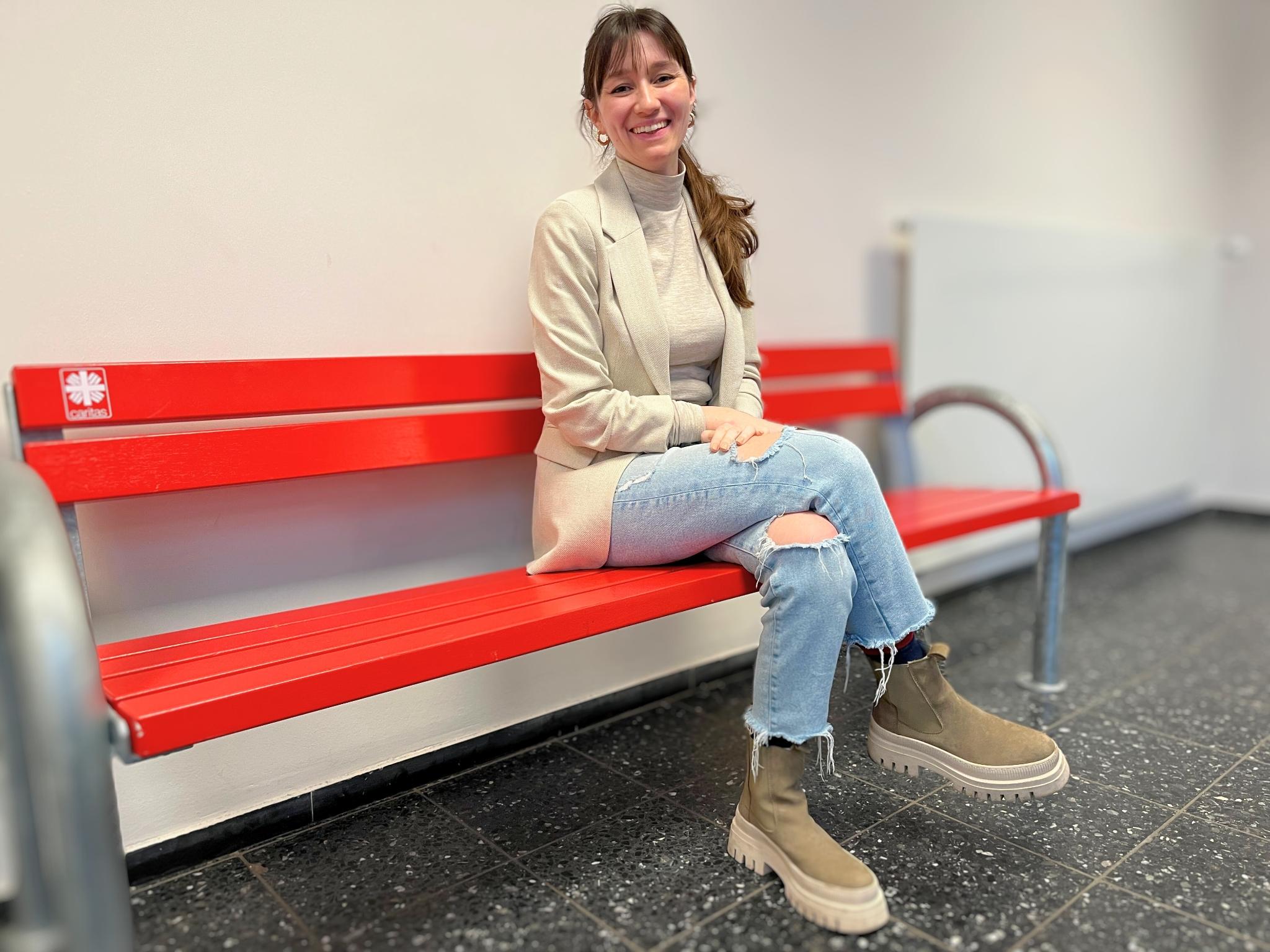 Auf dem Foto sitzt Hannah Matthews auf einer roten Bank mit einem kleinen Caritas-Logo. Sie trägt Jeans, einen hellen, beigefarbenen Pulli und eine Jacke in fast gleicher Farbe. Der Kopf ist nach rechts zur Kamera gewendet, die langen braunen Haare fallen in einem Pferdeschwanz über die linke Schulter, sie lächelt.