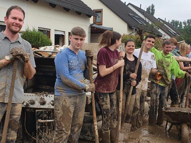 Das Foto wurde im Ahrtal aufgenommen zeigt eine verschlammte Straße, im Hintergrund Ein-Familien-Häuser, rechts am Bildrand ein Radlader. Im Mittelpunkt etwa ein Dutzend Menschen, die in einer Reihe nebeneinanderstehen. Alle haben Schaufeln in der Hand, sind schlammverdreckt, wirken erschöpft, lächeln aber; es handelt sich wohl um freiwillige Helfer.