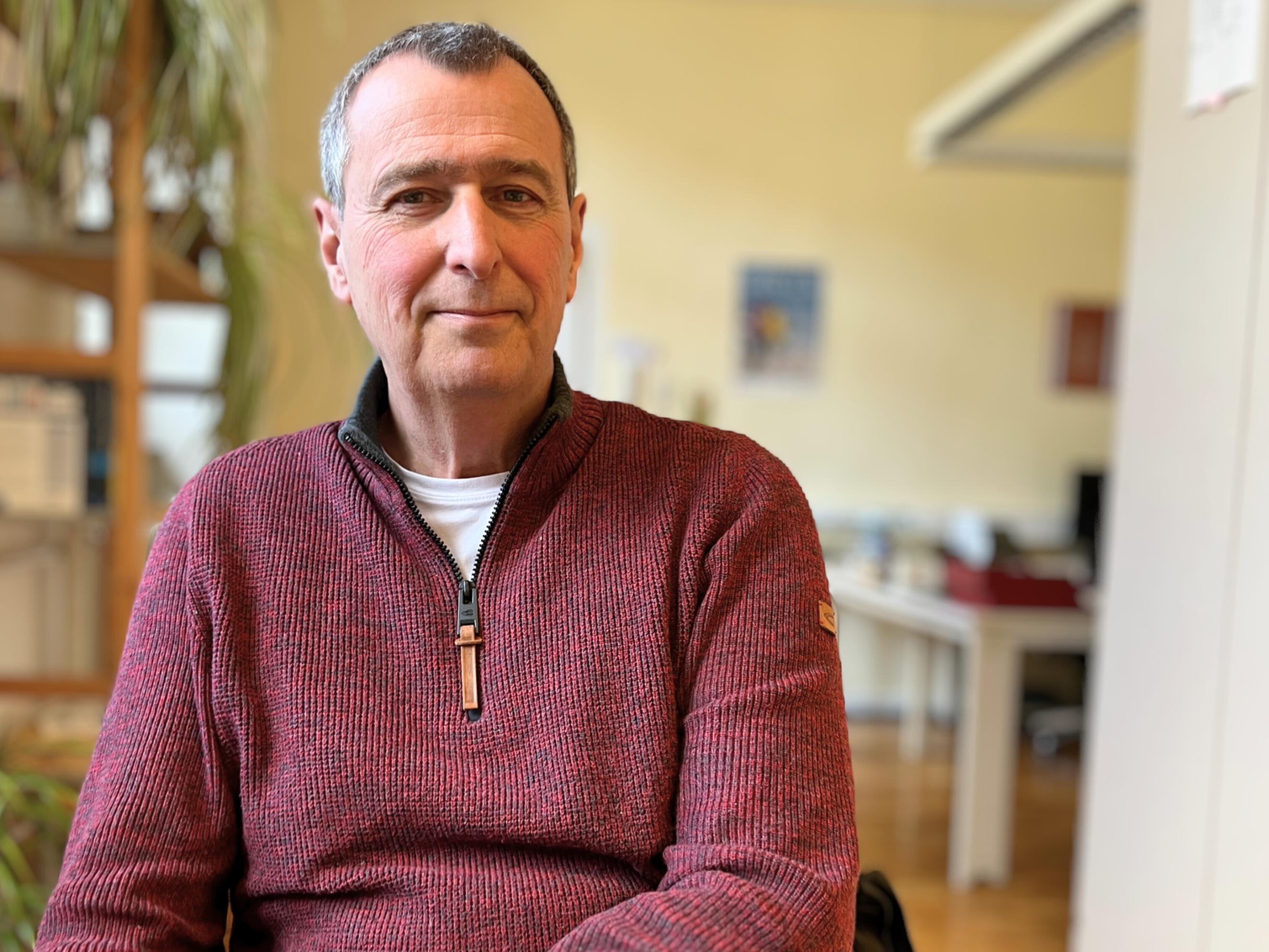 Ein älterer Mann (es handelt sich um Hubertus Kesselheim) mit kurzen grauen Haaren und freundlichem Gesichtsausdruck sitzt in einem Büro und schaut direkt in die Kamera. Er trägt einen rotbraunen Strickpullover mit Reißverschluss über einem weißen T-Shirt. Im Hintergrund sind unscharf ein Schreibtisch, Pflanzen und Büromaterialien zu sehen. (Alt-Text erstellt mit Unterstützung von KI)