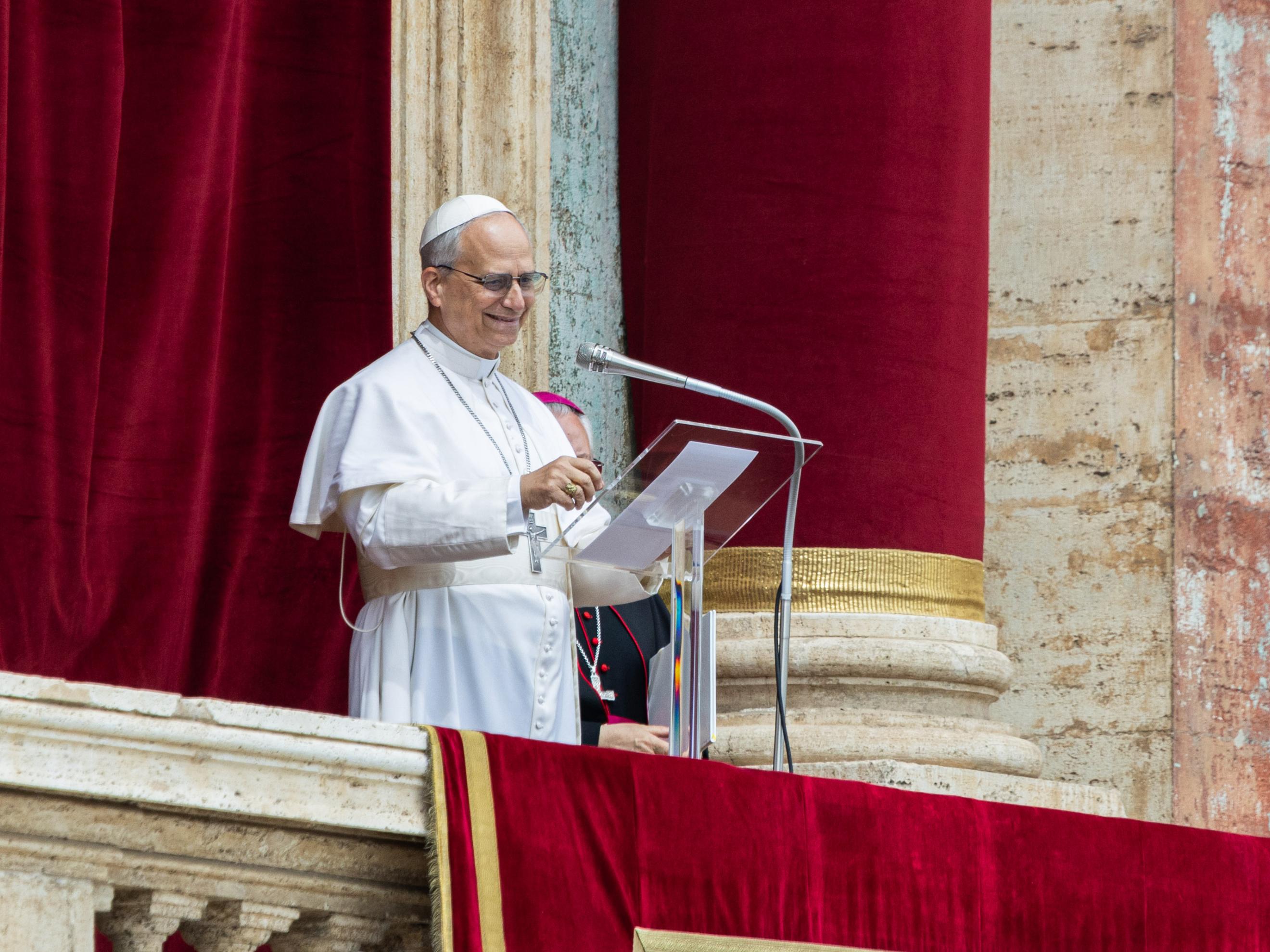 Papst Leo XIV. steht in weißer päpstlicher Kleidung auf der Loggia des Petersdoms vor einem roten Samtvorhang und spricht an einem Mikrofon. Er trägt ein Brustkreuz und eine weiße Kappe (Zucchetto) und lächelt. Eine weitere geistliche Person in dunkler Kleidung ist teilweise hinter ihm zu sehen.