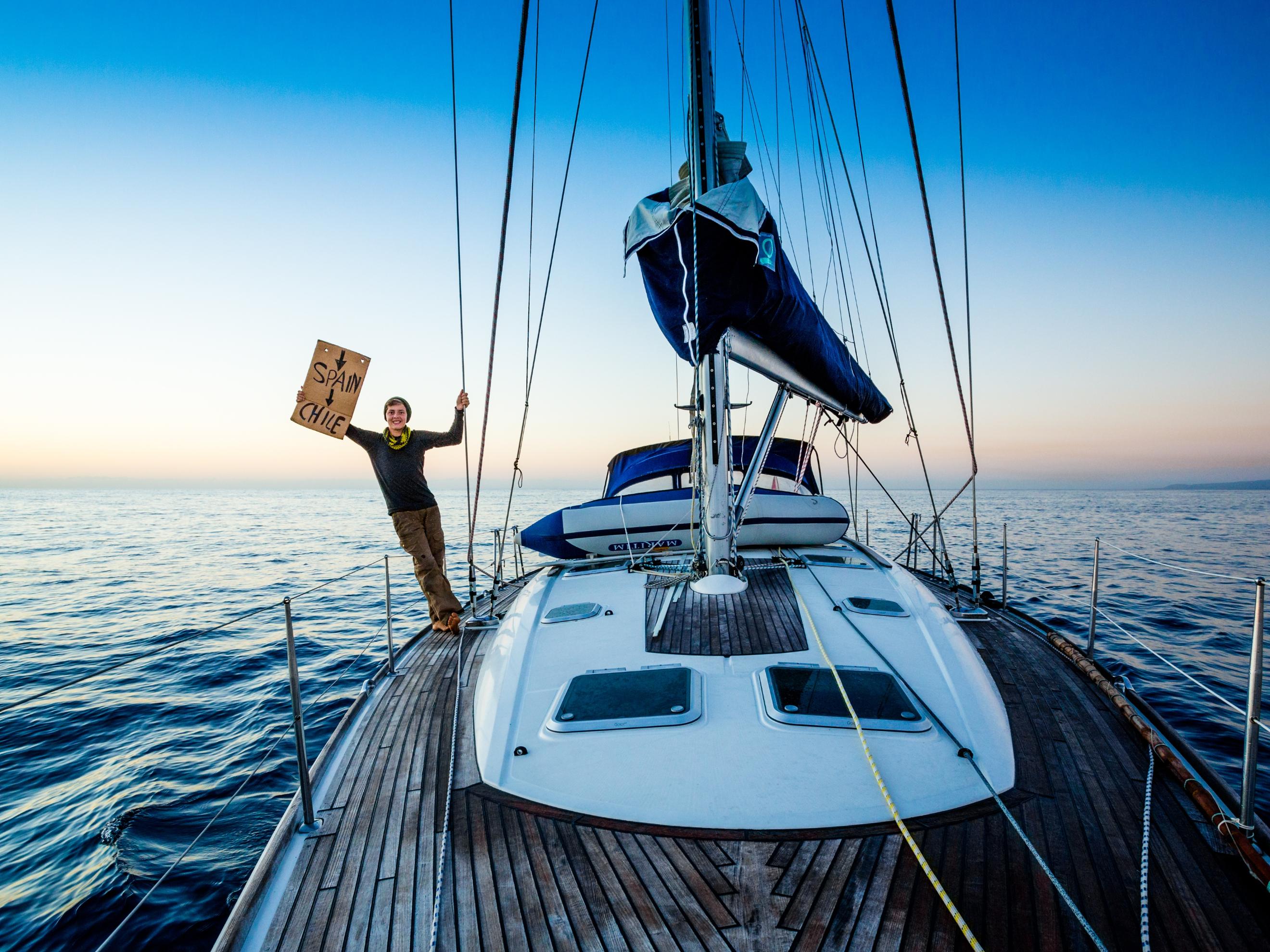 Joshi Nichell steht auf einem weißen Segelboot, das sich auf offener See befindet. Mit einer Hand hält er sich am Segel fest und lehnt sich über das Boot hinaus; mit der anderen Hand hält er ein Schild fest.