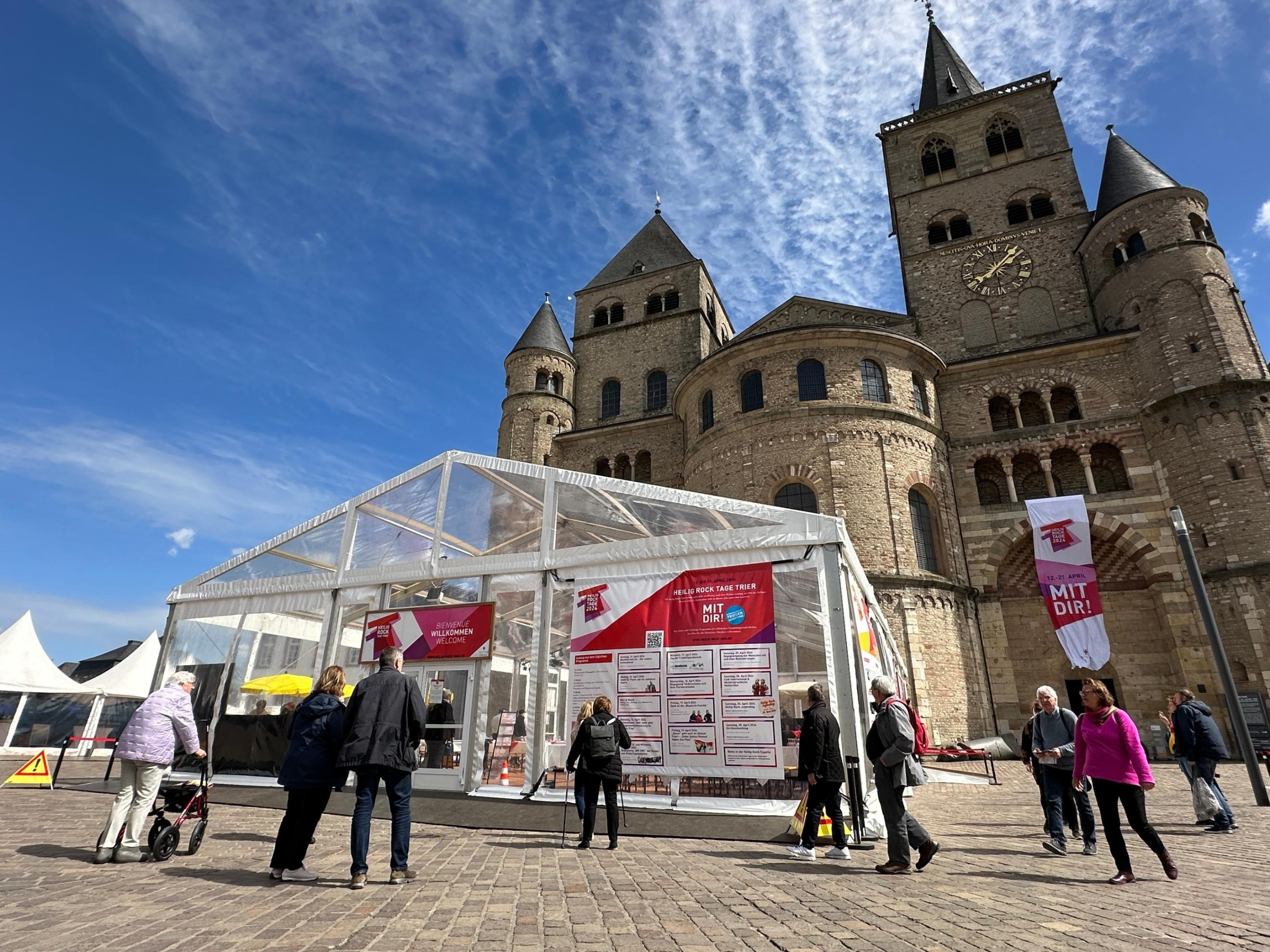 Man sieht das Kulturzelt vor dem Dom, der Himmel ist blau; im Vordegrund stehen einige Menschen auf dem Domfreihof-