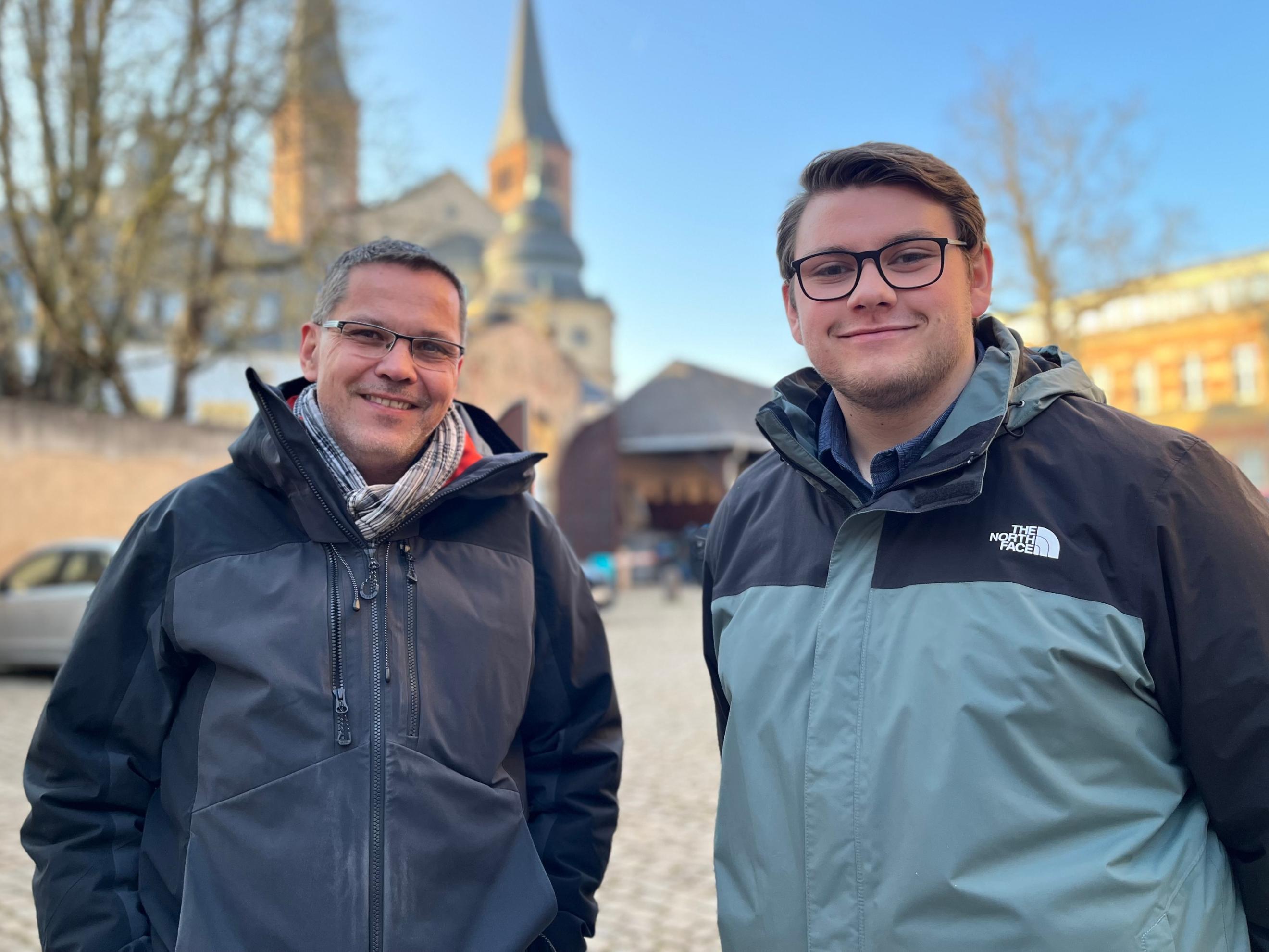 Man sieht zwei Männer auf einem Parkplatz, die in die Kamera lächeln. Im Hintergrund ist unscharf der Trierer Dom zu sehen