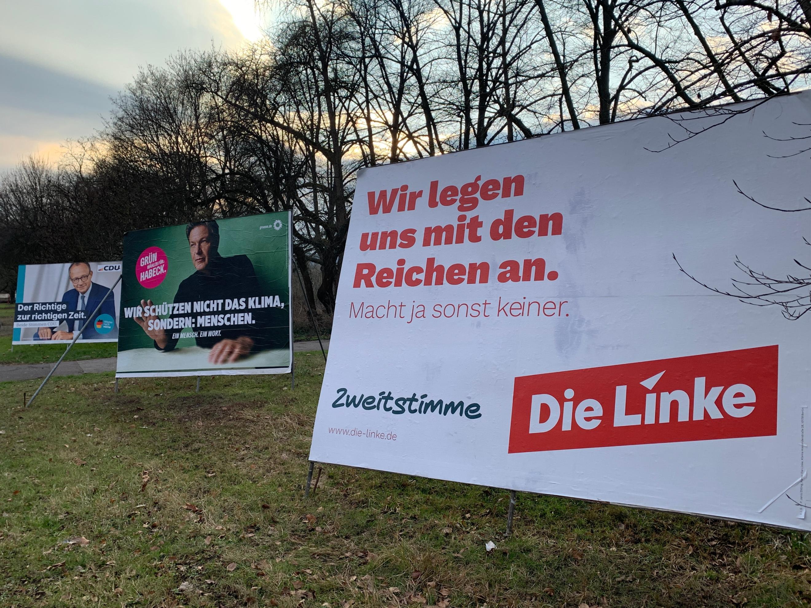 Das Bild zeigt drei große Wahlplakate verschiedener Parteien auf einer Wiese mit kahlen Bäumen im Hintergrund. Im Vordergrund steht ein Plakat der Partei Die Linke mit der Aufschrift „Wir legen uns mit den Reichen an.“ Daneben sind Plakate der Grünen mit Robert Habeck und der CDU mit Friedrich Merz zu sehen, jeweils mit ihren eigenen Wahlbotschaften.  (Alt-Text erstellt mit ChatGPT)