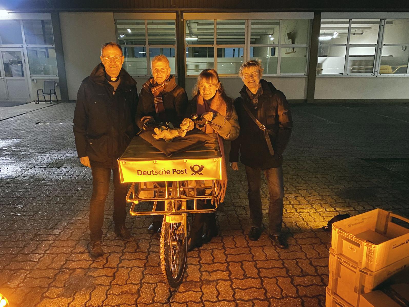Pfarrer Jörg Meyrer, Ulrike Phiesel, Nadine Kreuser und Bärbel Knieps vom Vor­bereitungsteam mit der Figur des Jesuskinds, das von einem Postboten per Fahrrad geliefert wurde.