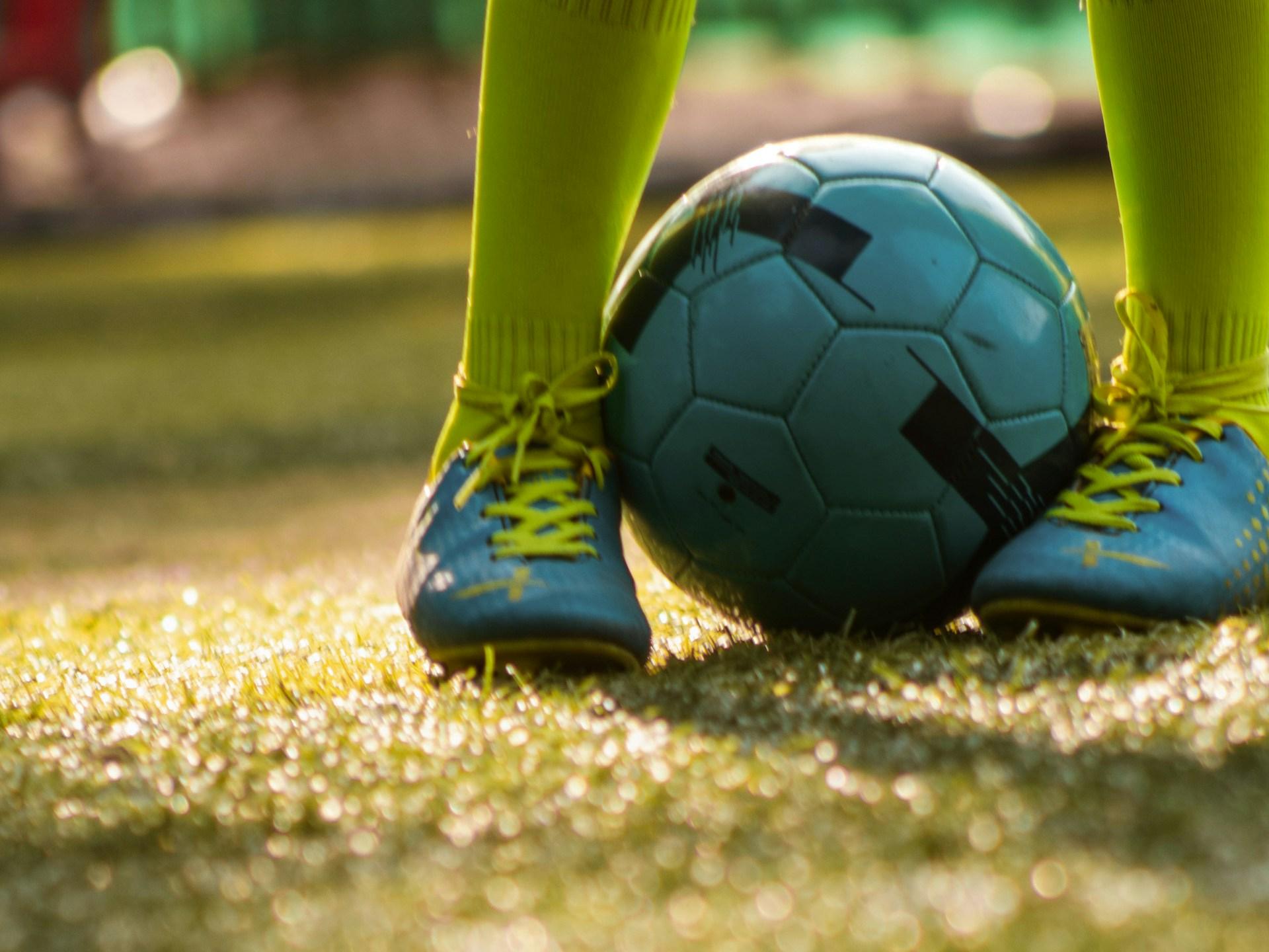 Das Foto zeigt auf einer Wiese in Nahaufnahme zwei Kinderfüße in blauen Turnschuhen und gelb-grünen Strümpfen, die einen Fußball festhalten.