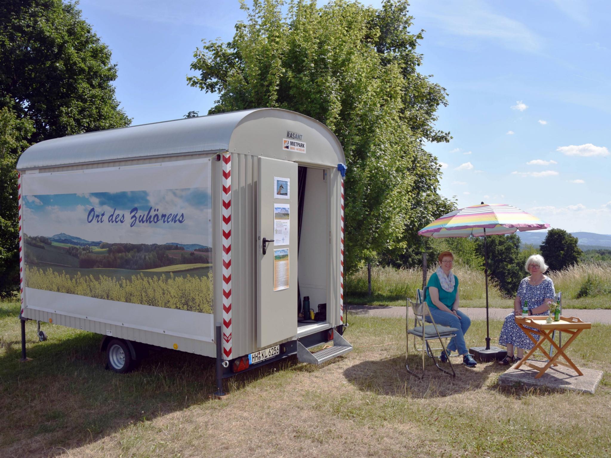 Pastoralreferentin Carola Fleck (links) und Ehrenamtliche Ute Morbach sitzen vor dem Bauwagen am Herzweg-Parkplatz. Foto: Ute Kirch/Bistum Trier