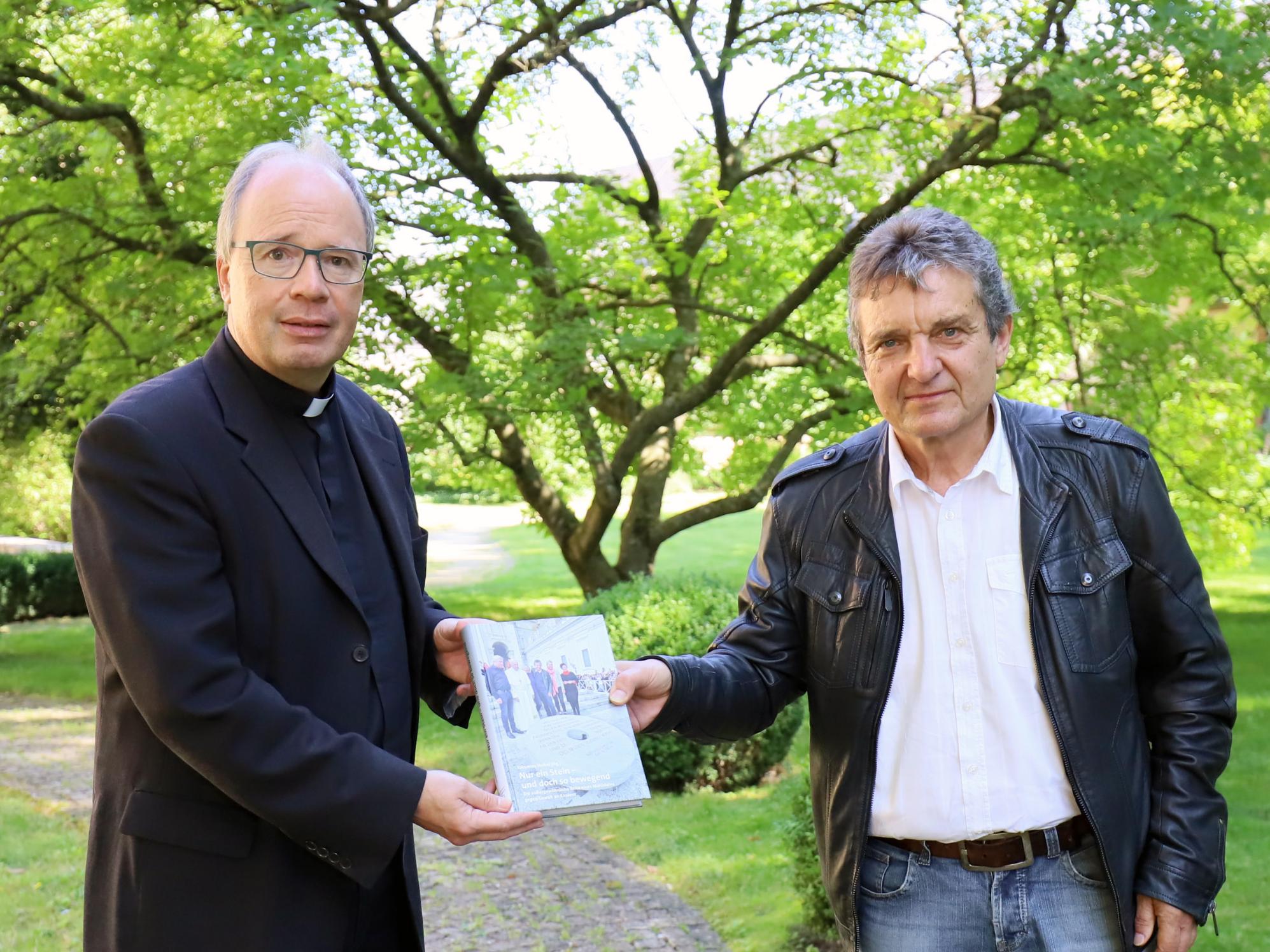 Johannes Heibel (links) übergibt den Bildband zum „Mahnenden Mühlstein“ an Bischof Ackermann. (Foto: Christina Libeaux/Bistum Trier)