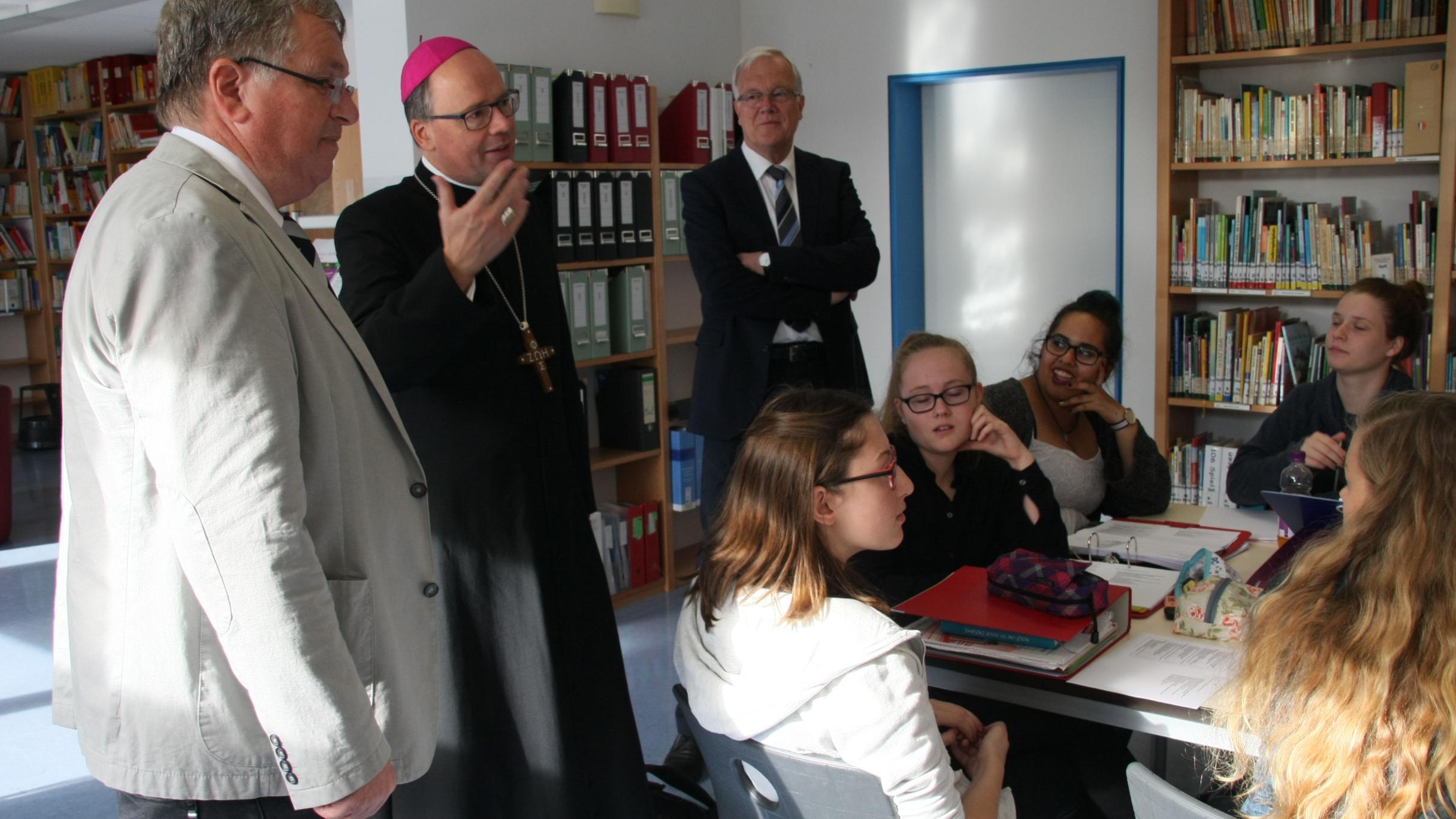 In der Bibliothek treffen Bischof Dr. Stephan Ackermann (Mitte), Wolfgang Müller vom Bischöflichen Generalvikariat (rechts) und Schulleiter Thomas Schmitz auf eine Gruppe von Schülerinnen, die sich auf ein anstehendes Referat vorbereiten.