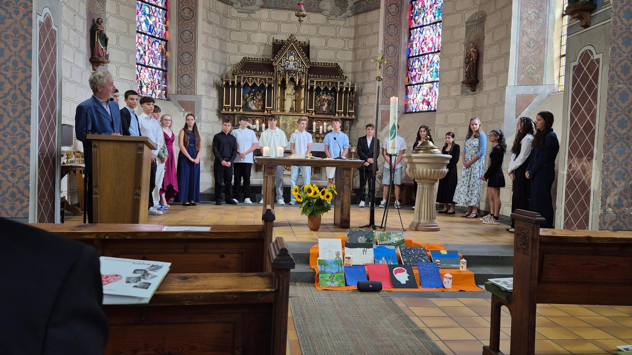 Das Bild zeigt eine Gruppe junger Menschen während der Firmung, die in einer Kirche vor einem Altar mit einem Blumenstrauß und bunten Tüchern stehen.