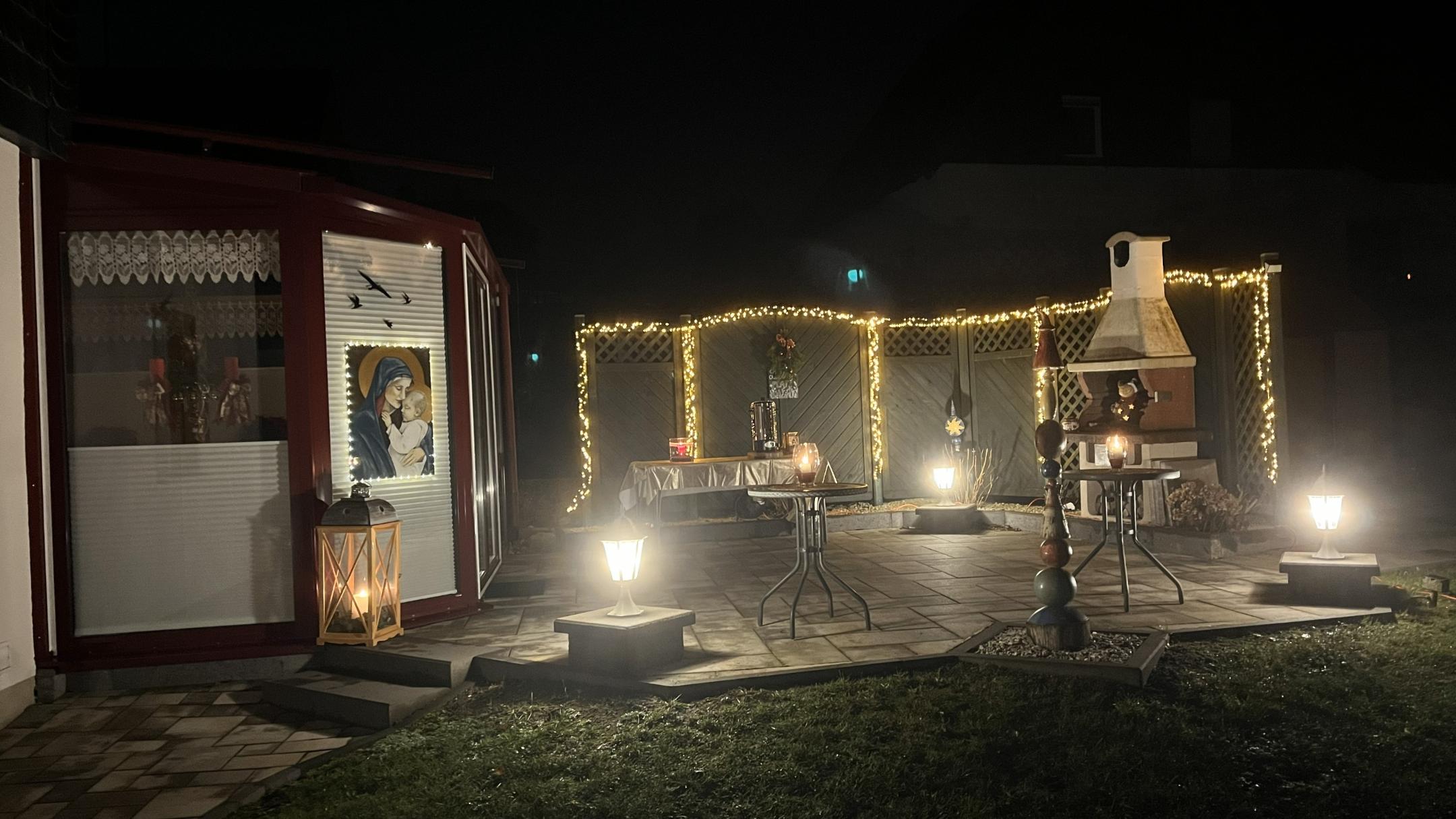 Die beleuchtete Terrasse mit Lichtern, Kerzen und dem Marienbild, Tischen und Kamin im Freien.