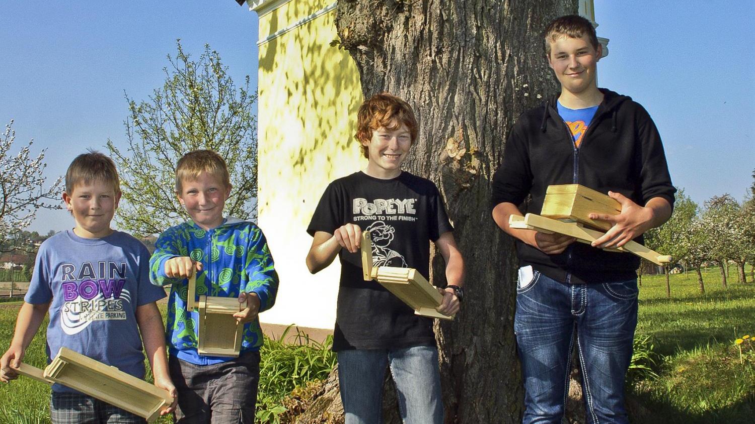 Vier Personen stehen im Freien vor einem Baum und halten selbstgebaute Holzobjekte in den Händen.