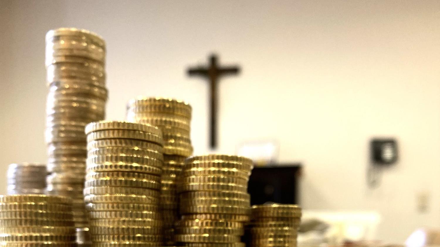 Auf dem Foto stehen viele gestapelte Münzen auf einem Tisch; im Hintergrund hängt ein schlichtes Kreuz an der Wand.