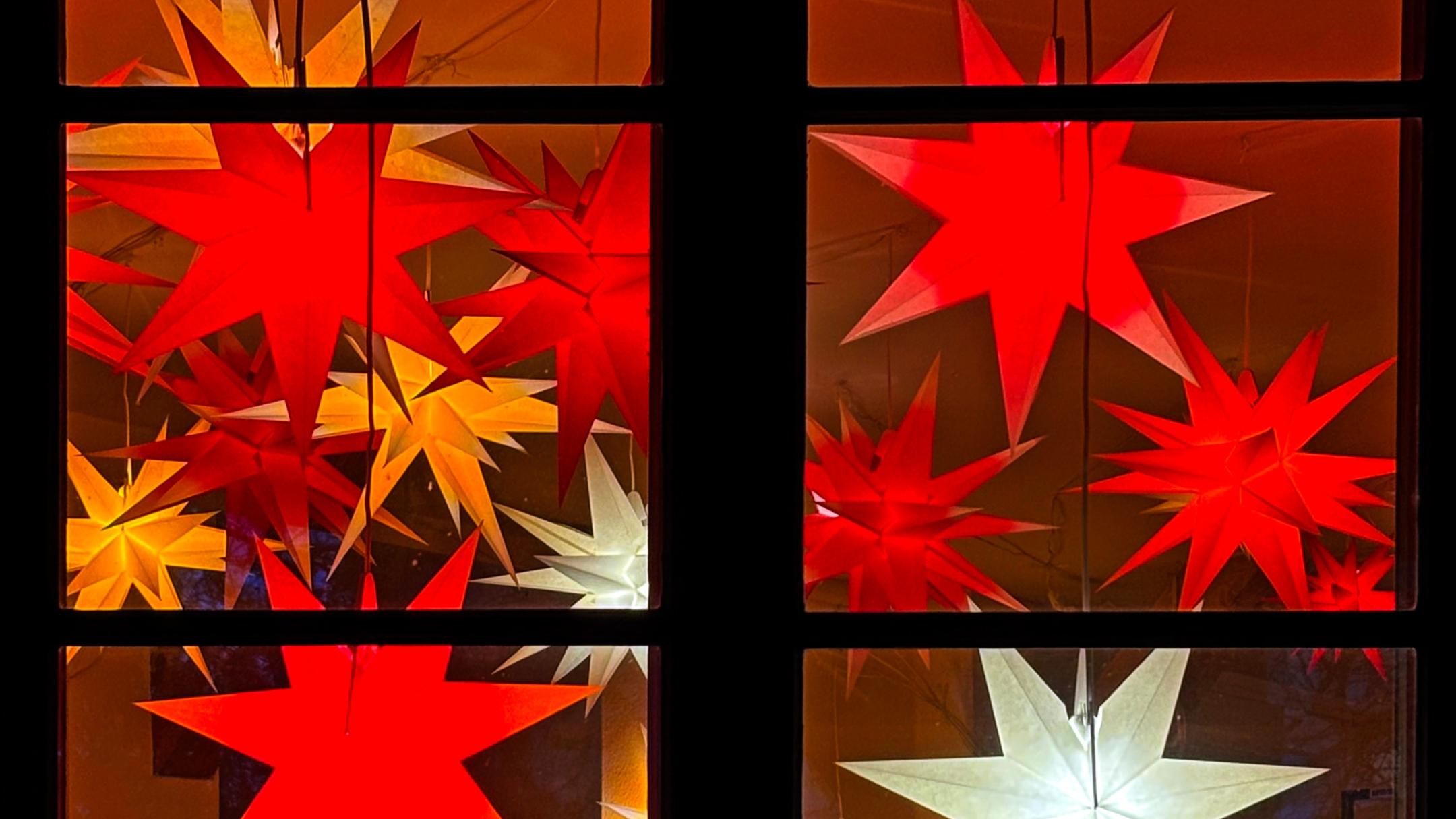Das Bild zeigt ein Fenster, vor dem mehrere leuchtende Sterne in Rot, Gelb und Weiß an Schnüren hängen und eine warme, festliche Stimmung erzeugen.