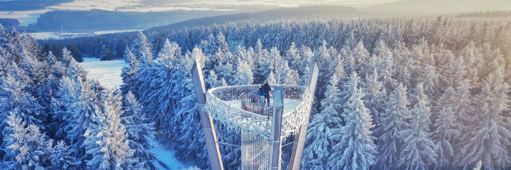 Zu sehen ist ein Metallturm mit Platzform, auf der zwei Menschen stehen. Rund herum stehen verschneite Bäume, im Hintergrund ist der Himmel leicht bewölkt. Die Aufnahme wurde mit einer Drohne von oben gemacht