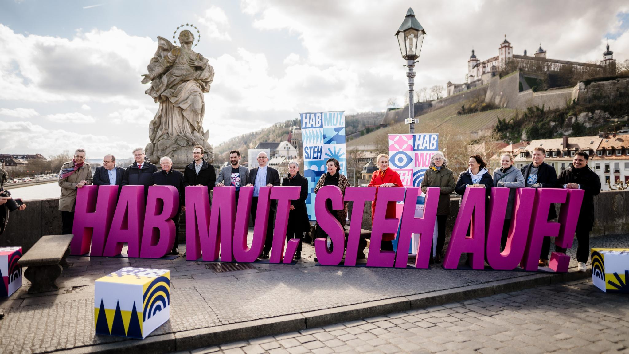 15 Menschen halten auf der Alten Mainbrücke die violetten Einzel-Großbuchstaben des Katholikentagsmottos: 'Hab Mut, steh auf!'