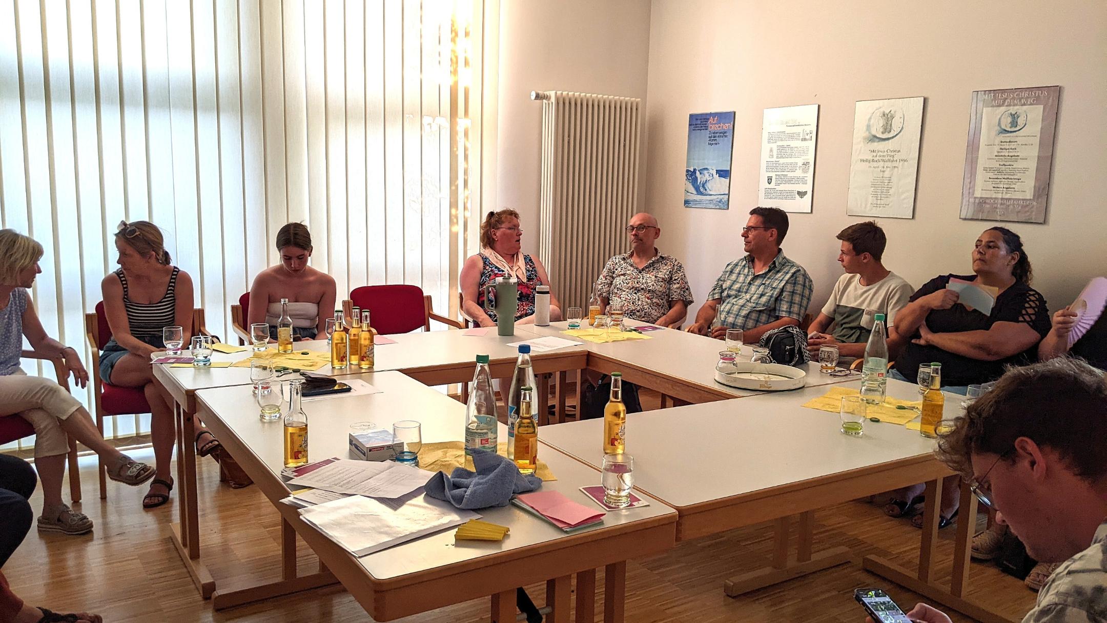 Mehrere Personen sitzen zusammen an Tischen in einem Viereck angeordnet und reden miteinander