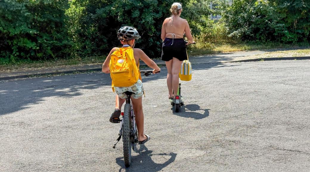Ein Kind mir gelbem Rucksack fährt Fahrrad, davor geht eine Frau