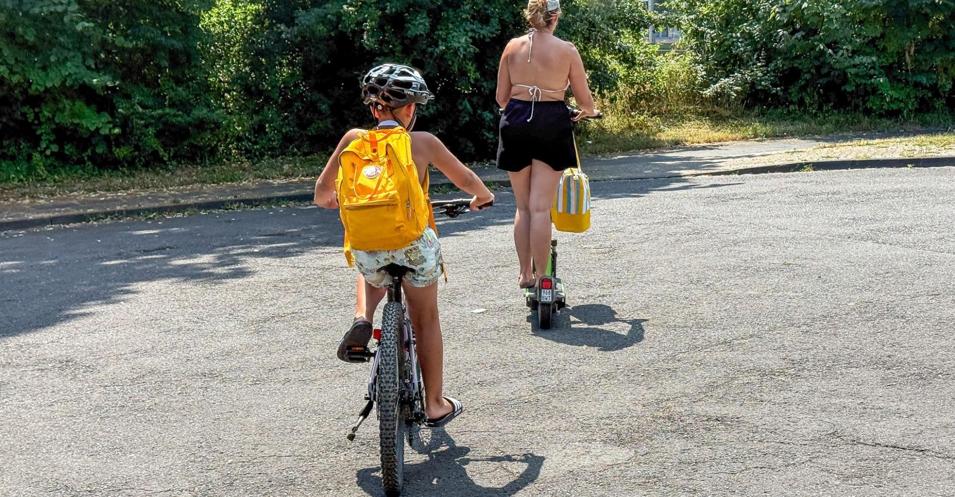 Ein Kind mir gelbem Rucksack fährt Fahrrad, davor geht eine Frau