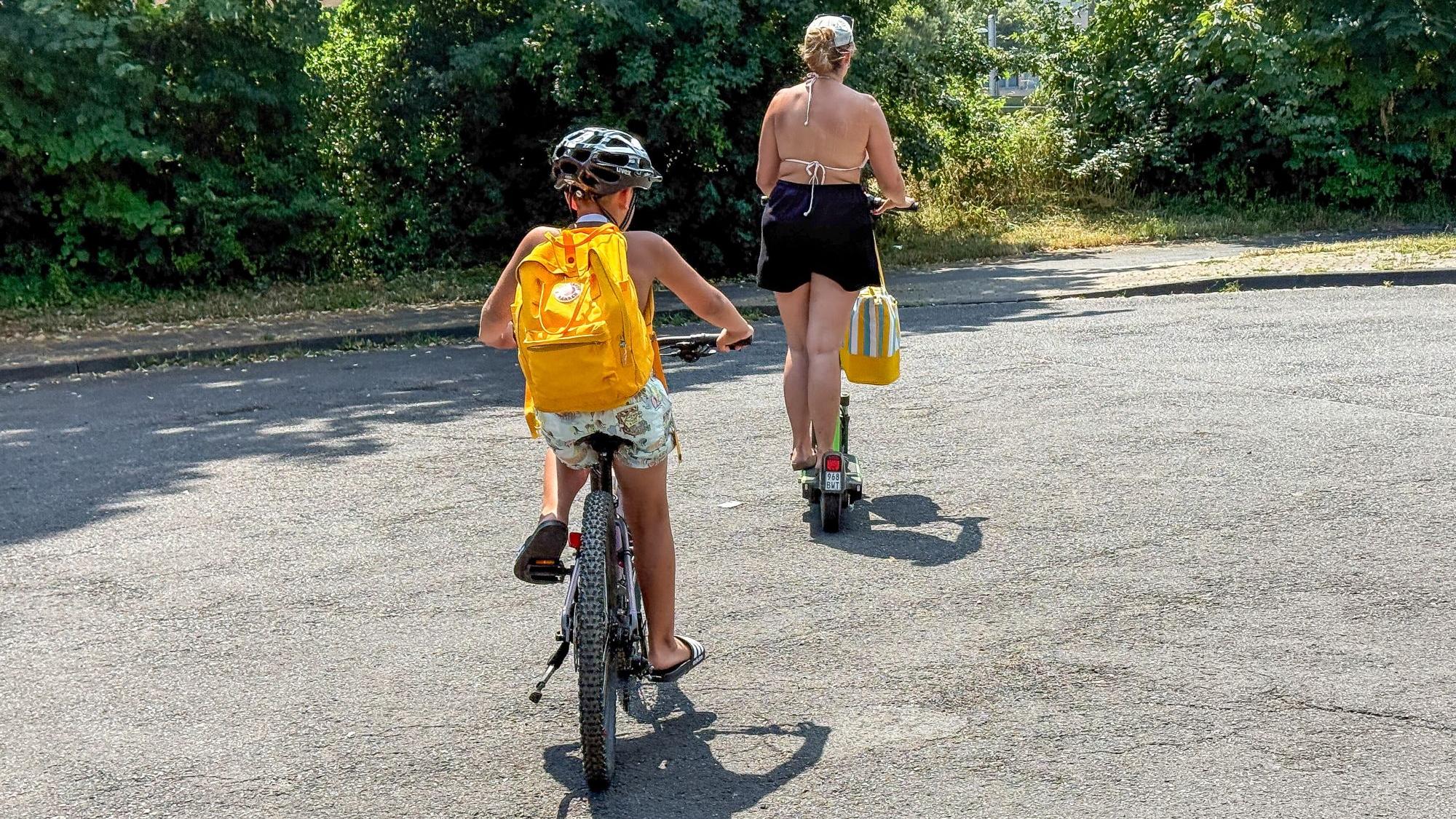 Ein Kind mir gelbem Rucksack fährt Fahrrad, davor geht eine Frau
