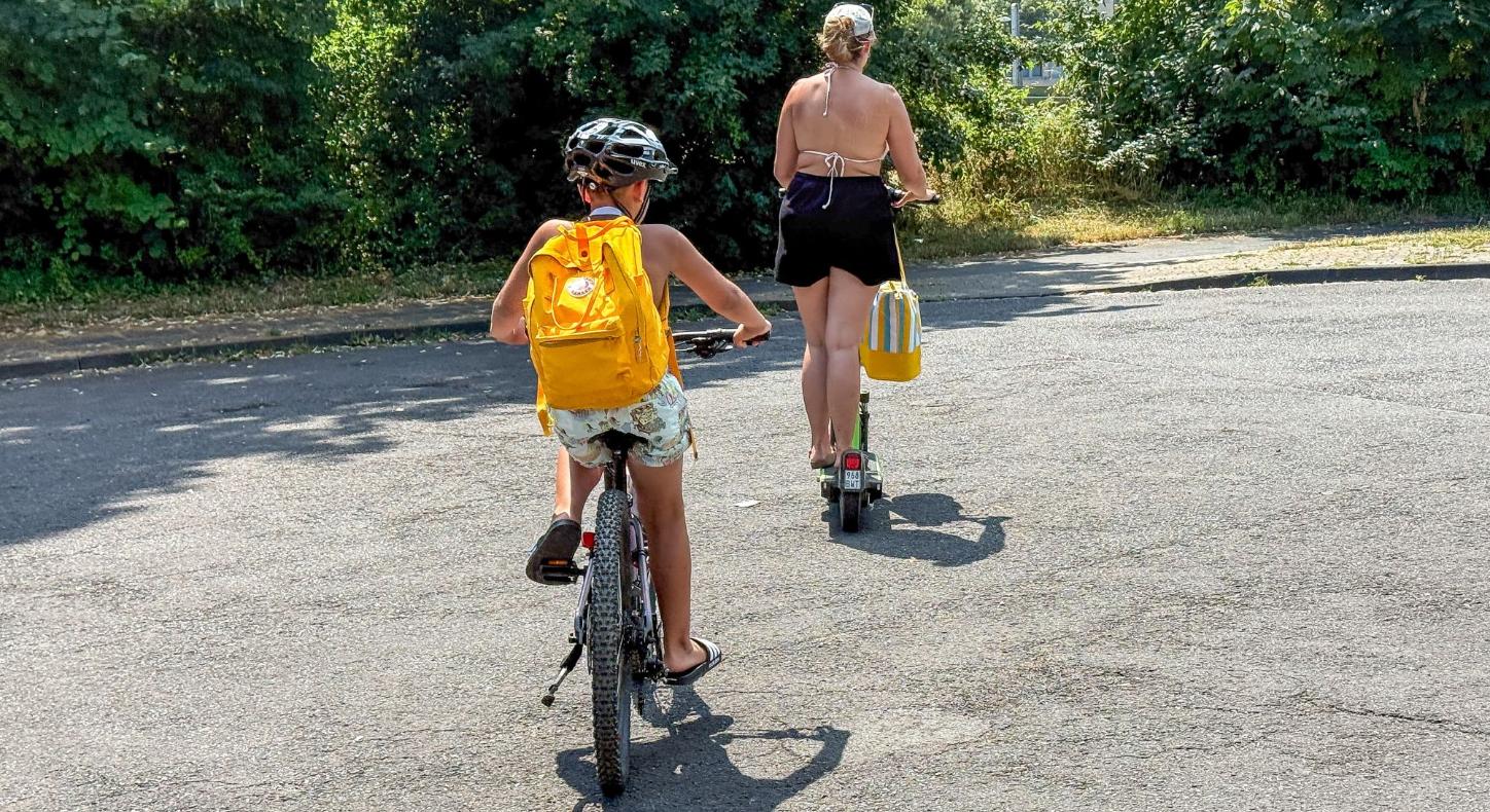 Ein Kind mir gelbem Rucksack fährt Fahrrad, davor geht eine Frau