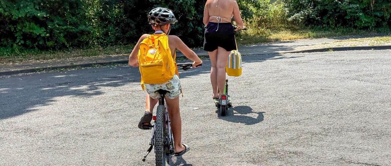 Ein Kind mir gelbem Rucksack fährt Fahrrad, davor geht eine Frau