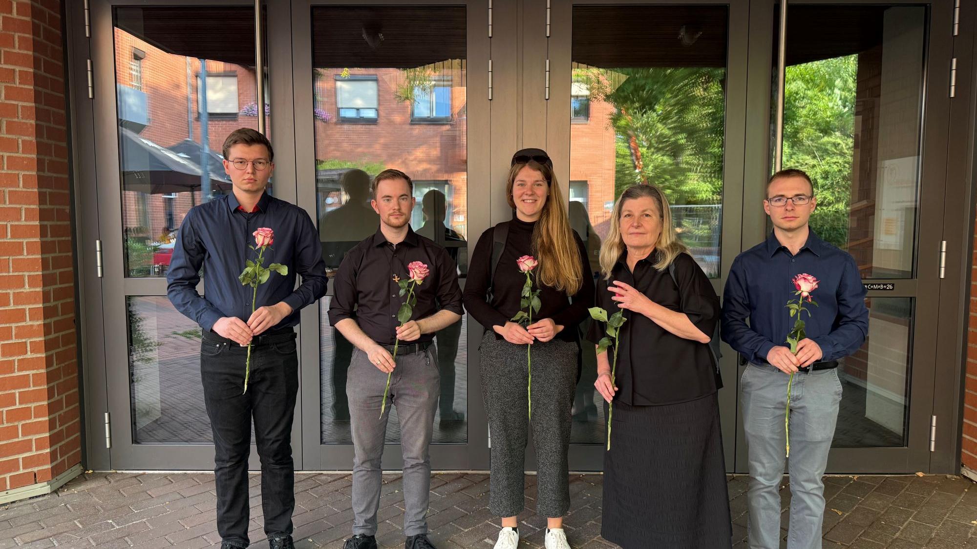 Dunkel gekleidete Menschen blicken in die Kamera und halten Rosen in den Händen