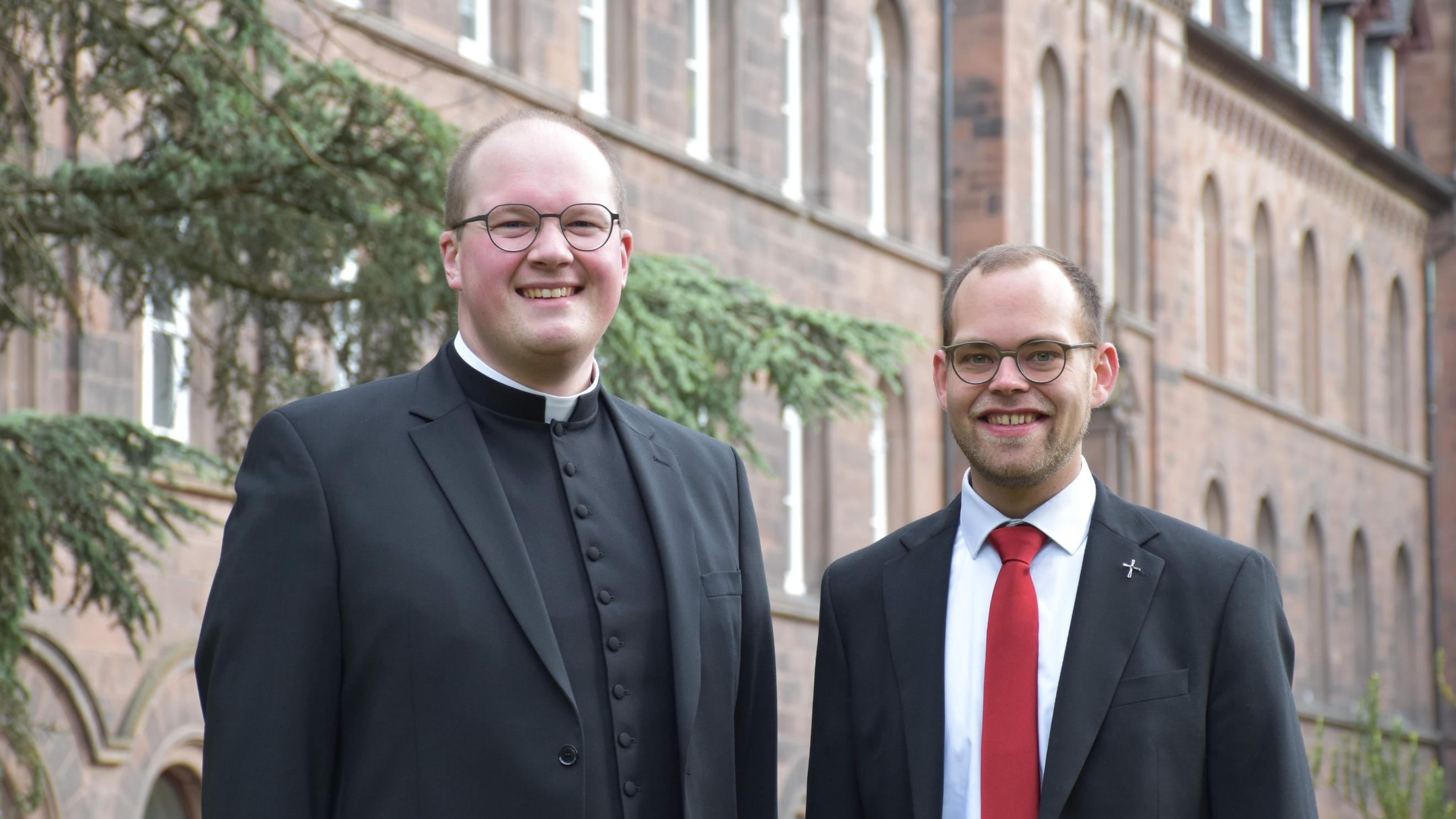 Zwei Männer vor einem rötlichen Gebäude lächeln in die Kamera. Einer trägt einen priesterkragen, einer eine rote Krawatte