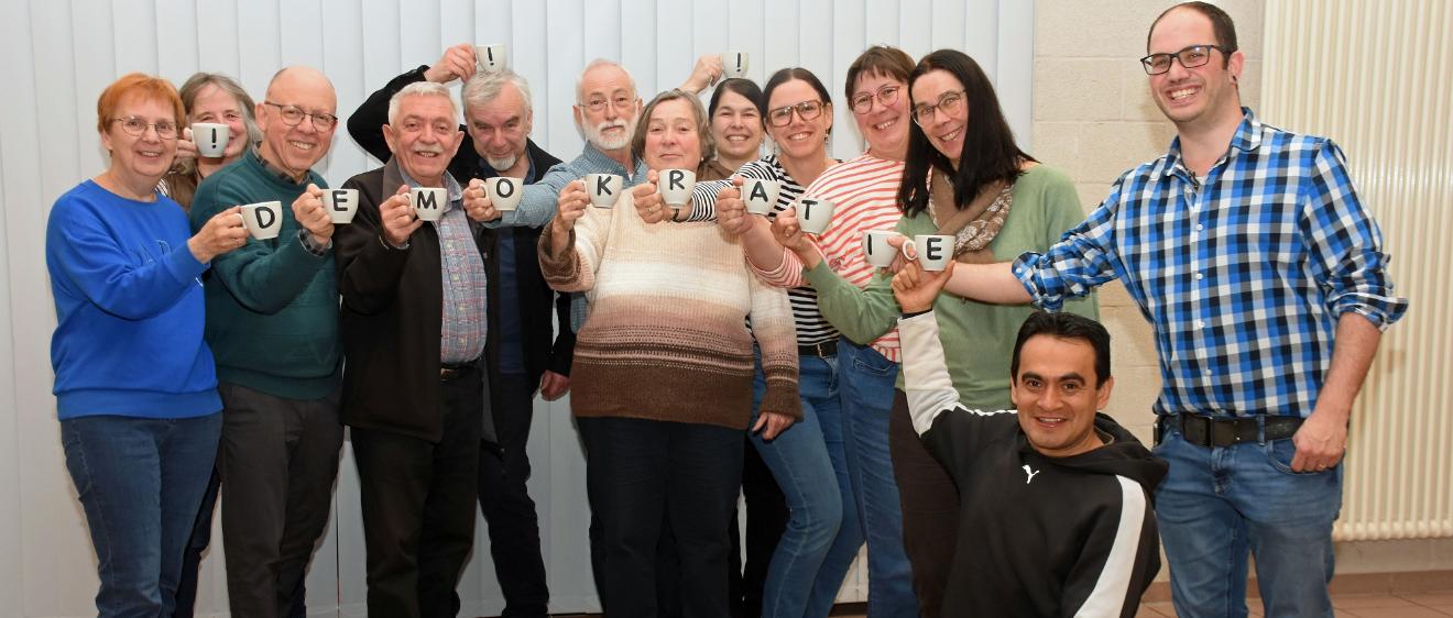 Die Mitglieder des Pfarrgemeinderates halten mit Buchstaben bedruckte Tassen, die zusammen das Wort Demokratie ergeben.