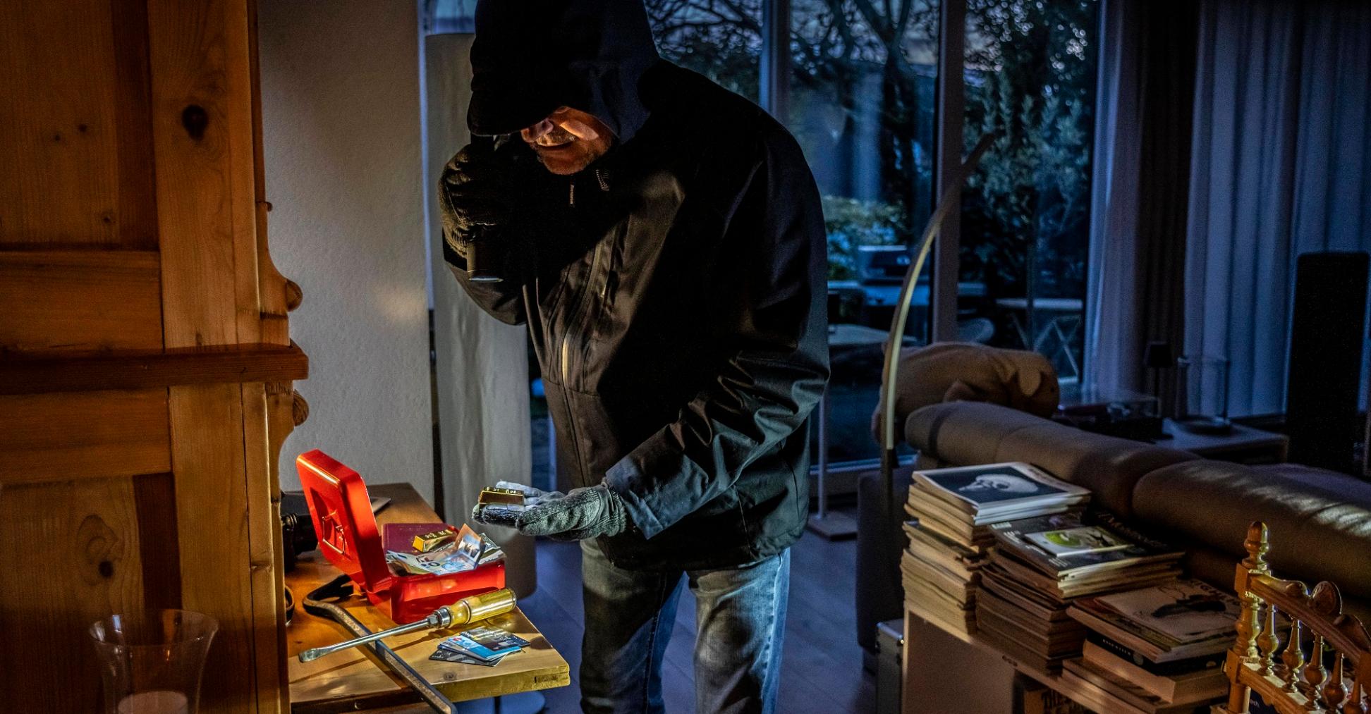 Ein dunkel gekleideter Mann mit Kapuze durchsucht mit einer Taschenlampe eine aufgebrochene Geldkassette.
