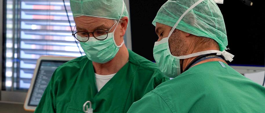 Zwei Männer in Grünen Hemden mit Mundschutz arbeiten an einem Patienten