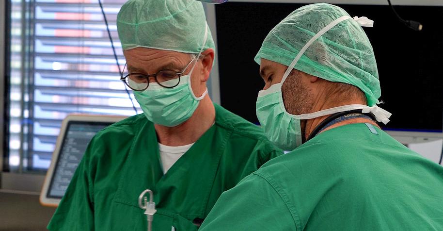 Zwei Männer in Grünen Hemden mit Mundschutz arbeiten an einem Patienten