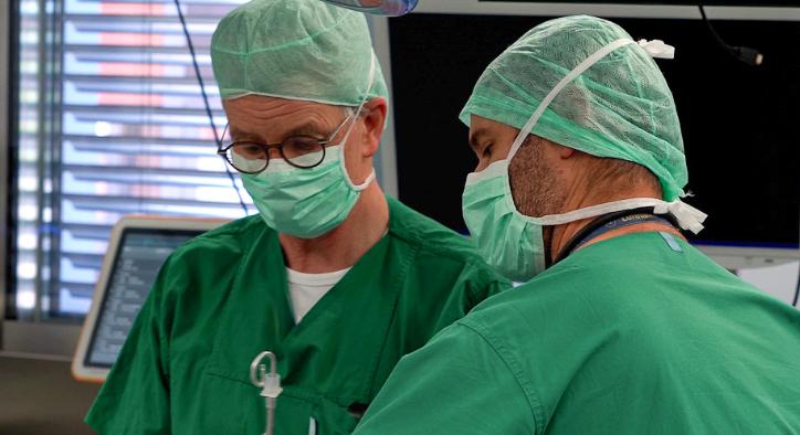 Zwei Männer in Grünen Hemden mit Mundschutz arbeiten an einem Patienten