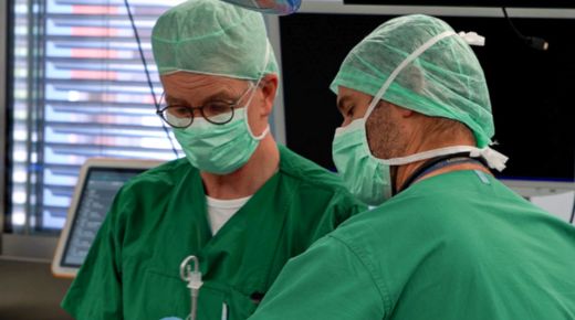 Zwei Männer in Grünen Hemden mit Mundschutz arbeiten an einem Patienten