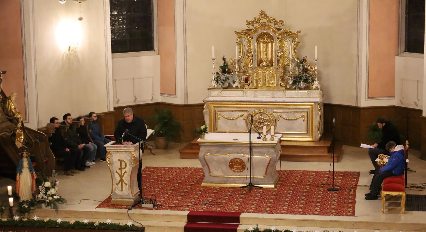 Altar von oben; ein Priester predigt; auf den Seitenbänken sitzen Personen und beten