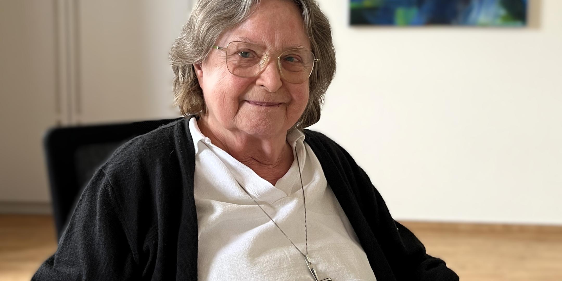 Sr. Evamaria Durchholz Ältere Frau mit grauen Haaren, Brille und einem großen silbernen Kreuz um den Hals sitzt an einem Tisch und lächelt in die Kamera