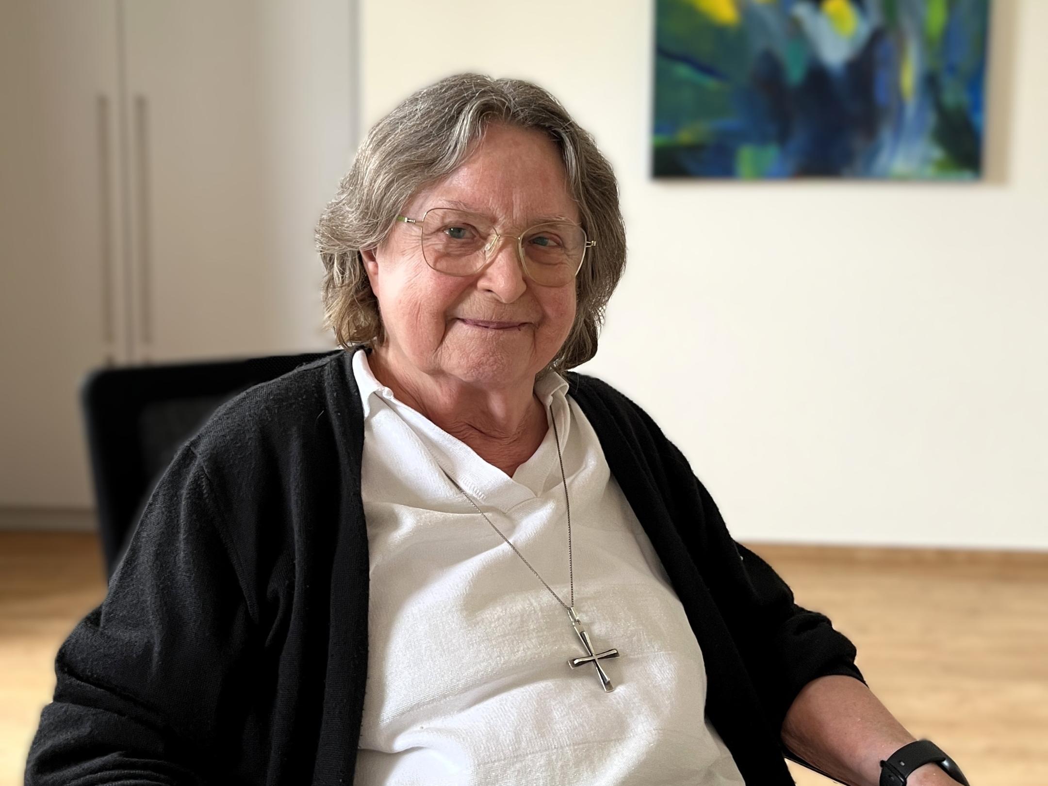 Ältere Frau mit grauen Haaren, Brille und einem großen silbernen Kreuz um den Hals sitzt an einem Tisch und lächelt in die Kamera