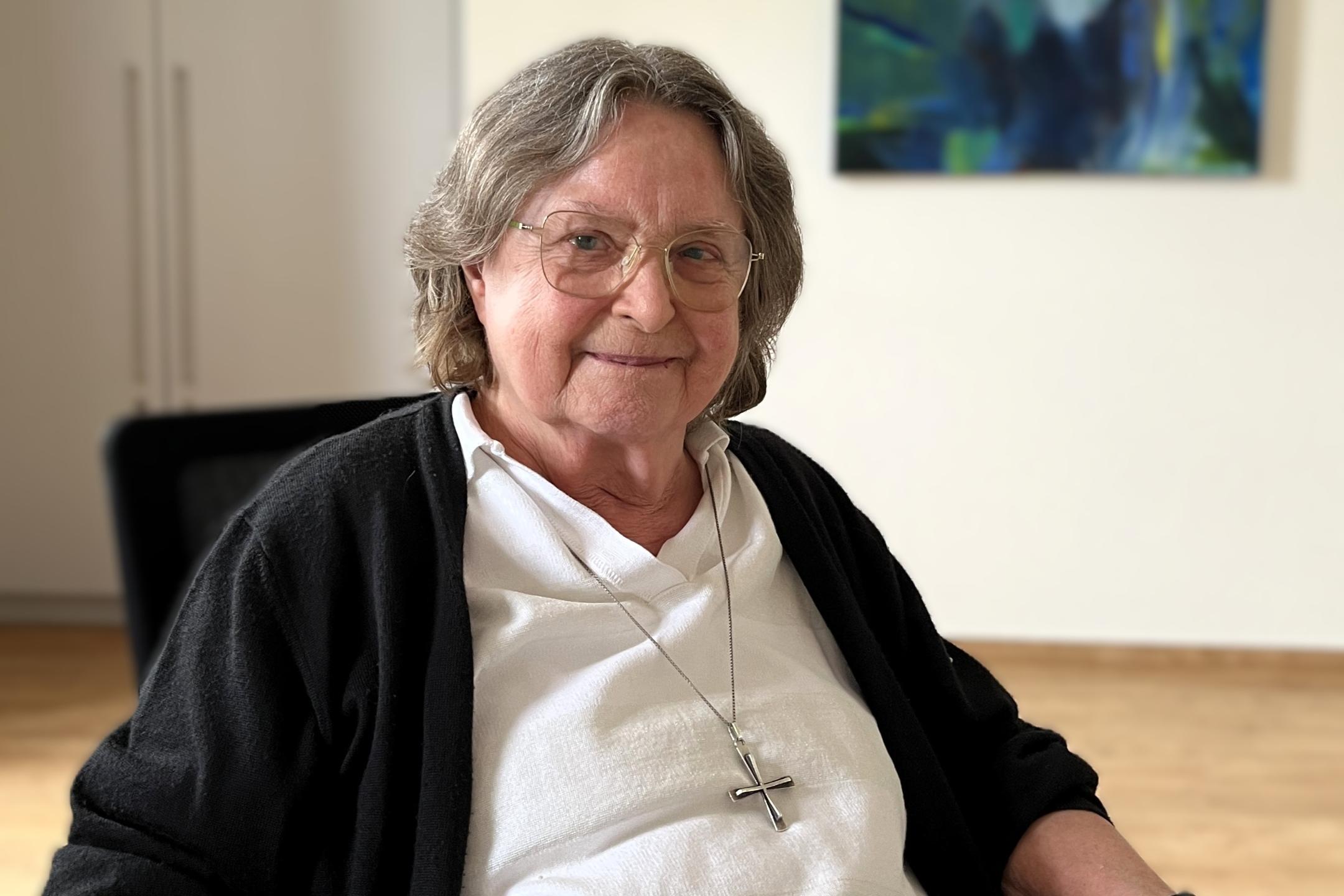 Ältere Frau mit grauen Haaren, Brille und einem großen silbernen Kreuz um den Hals sitzt an einem Tisch und lächelt in die Kamera