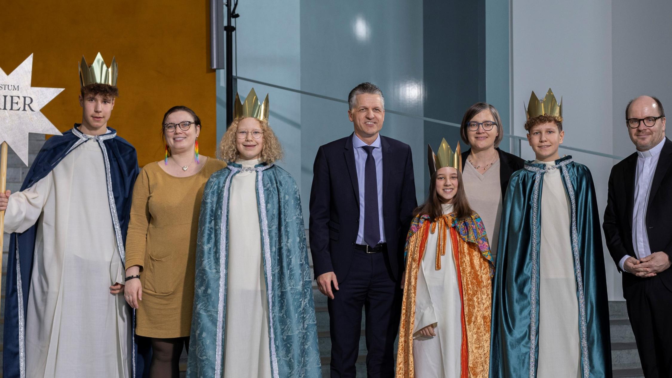 Vier Kinder mit Kronen und langen Samtumhängen stehen zwischen vier Erwachsenen; einer hält einen Stab mit einem Stern, auf dem Bistum Trier steht.