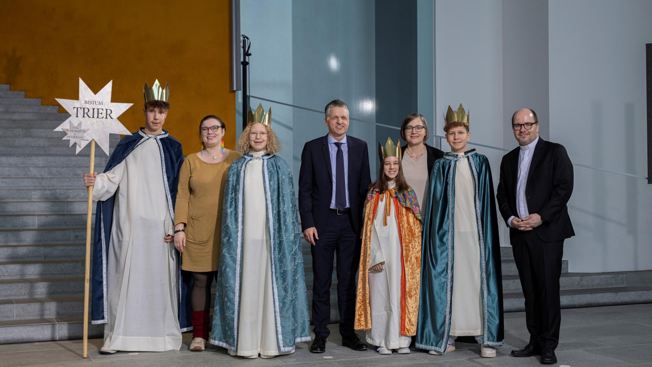 Vier Kinder mit Kronen und langen Samtumhängen stehen zwischen vier Erwachsenen; einer hält einen Stab mit einem Stern, auf dem Bistum Trier steht.