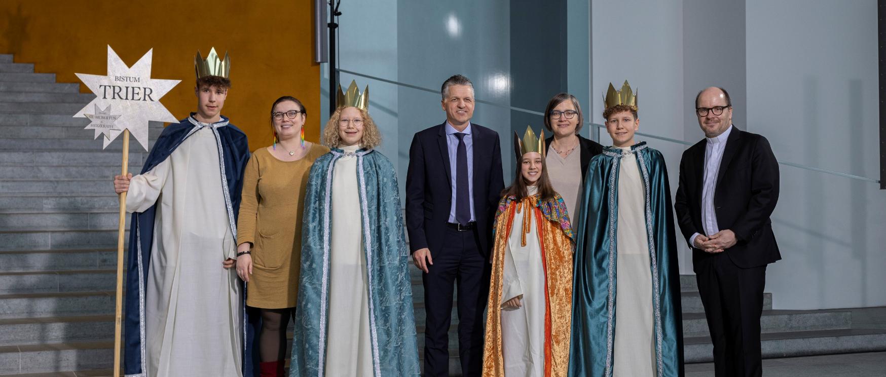 Vier Kinder mit Kronen und langen Samtumhängen stehen zwischen vier Erwachsenen; einer hält einen Stab mit einem Stern, auf dem Bistum Trier steht.