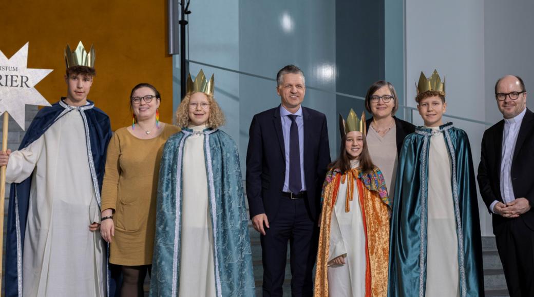 Vier Kinder mit Kronen und langen Samtumhängen stehen zwischen vier Erwachsenen; einer hält einen Stab mit einem Stern, auf dem Bistum Trier steht.