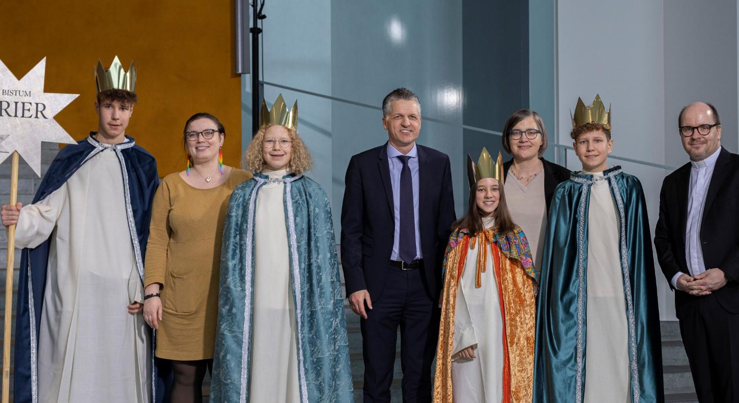 Vier Kinder mit Kronen und langen Samtumhängen stehen zwischen vier Erwachsenen; einer hält einen Stab mit einem Stern, auf dem Bistum Trier steht.